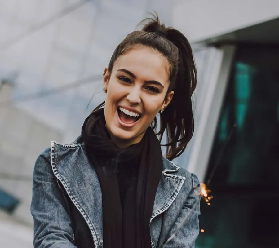 Smiling woman with ponytail wearing denim jacket and black scarf enjoying outdoor moment with sparkler
