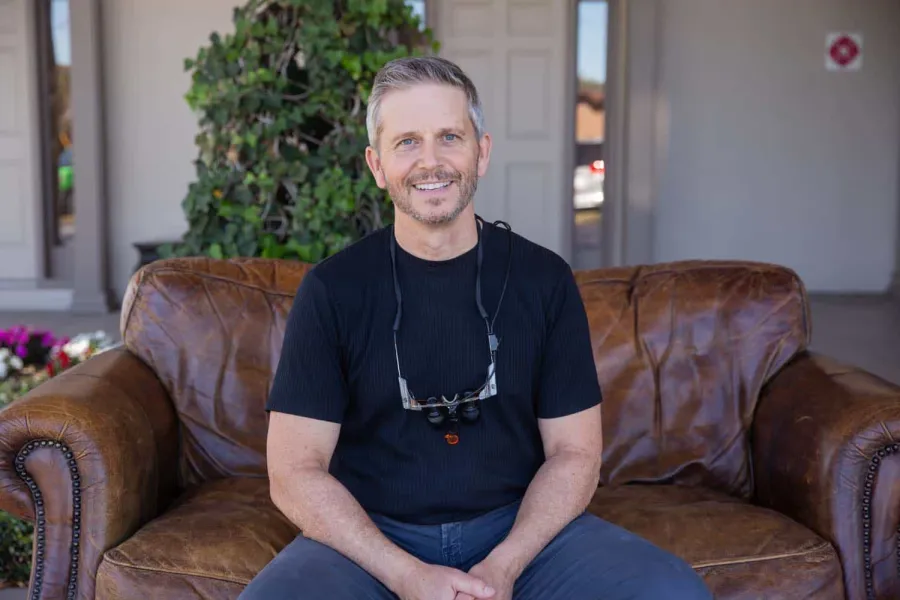 Smiling middle-aged man wearing black shirt and dental magnifying glasses sitting on a brown leather couch outdoors.
