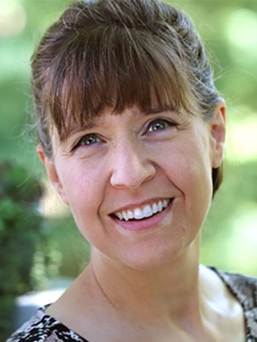 Smiling woman with brown hair and bangs in natural outdoor setting with green blurred background.