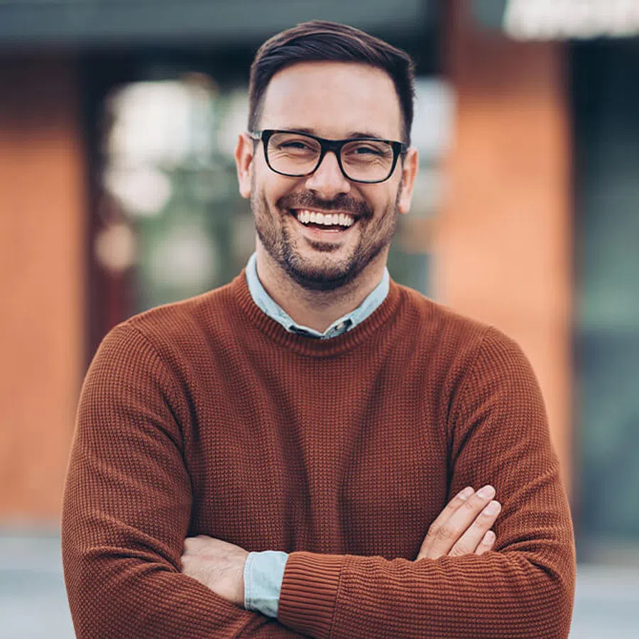 Smiling man with glasses and brown sweater standing outdoors with arms crossed