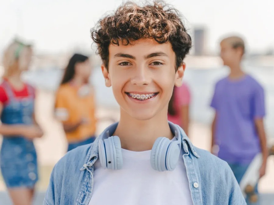 Smiling teenage boy with curly hair and braces wearing headphones around neck outdoors with blurred friends in background