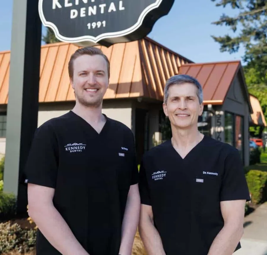 Two dentists wearing black Kennedy Dental uniforms stand outside the dental office under a Kennedy Dental sign.
