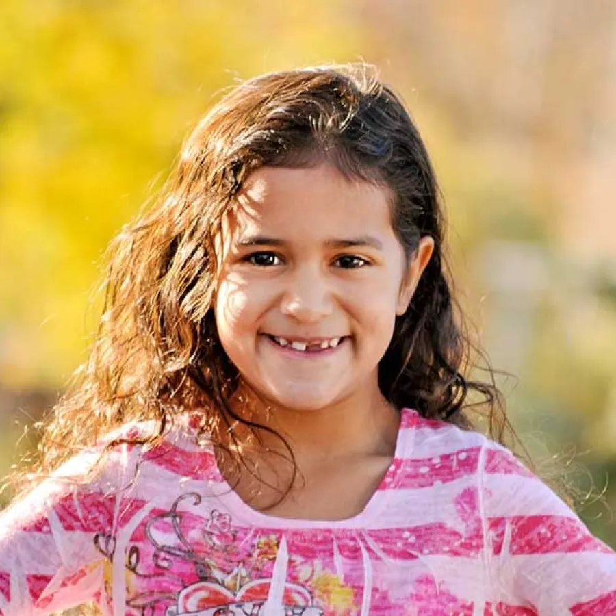 Smiling young girl with curly hair wearing a pink striped shirt outdoors with a blurred autumn background