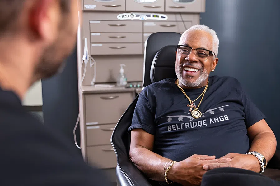 Smiling elderly man wearing glasses and jewelry sitting in a chair during a consultation in a modern office.