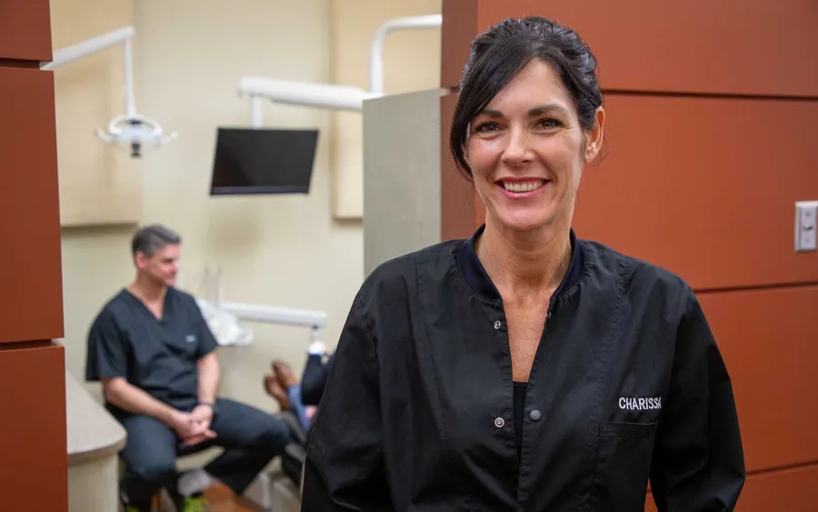Smiling female dental professional in black scrubs in a dental office with a colleague in the background
