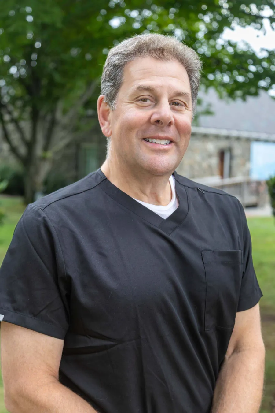 Middle-aged man wearing black medical scrubs smiling outdoors with green trees and a stone building in the background