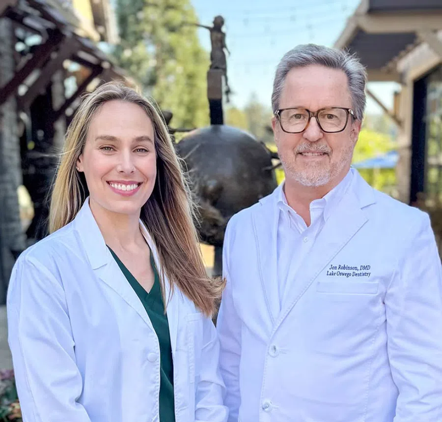 Two dental professionals in white coats standing outside smiling, with greenery and sculpture in background.