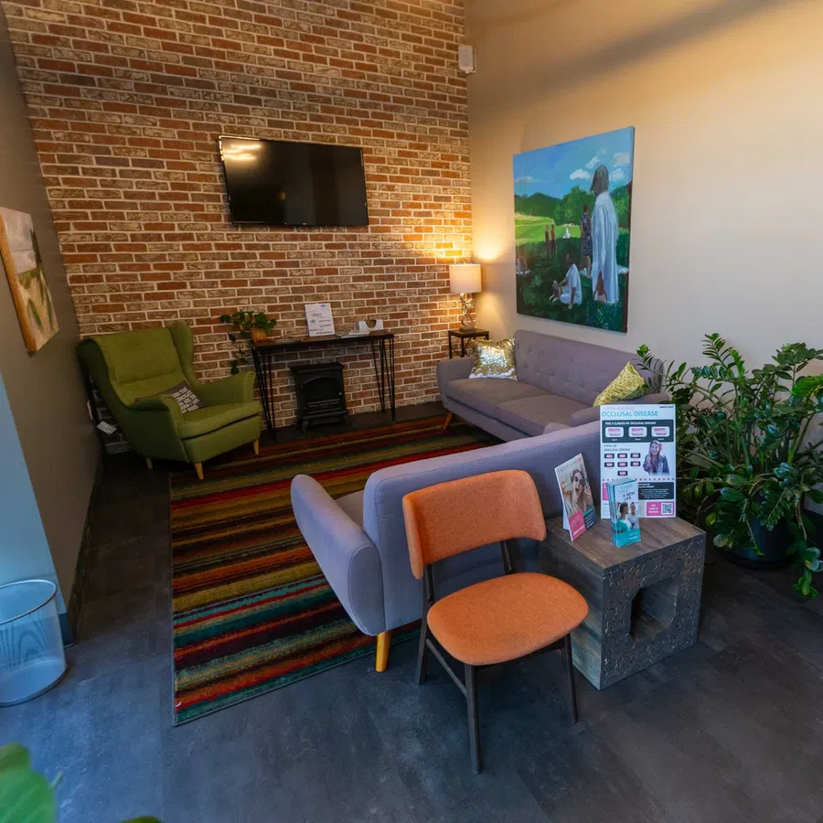 Cozy waiting room with brick wall, TV, green armchair, gray sofa, colorful rug, and indoor plants