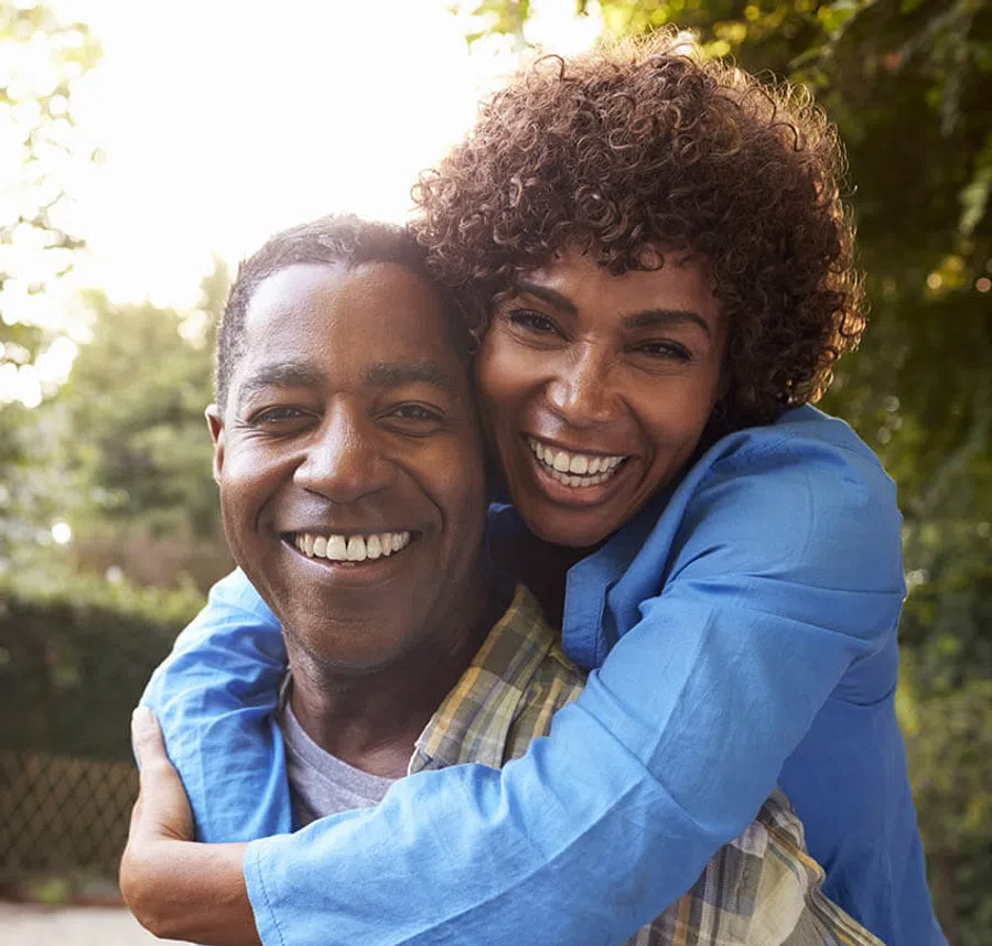 Happy couple embracing outdoors with bright smiles and natural background of trees and sunlight.