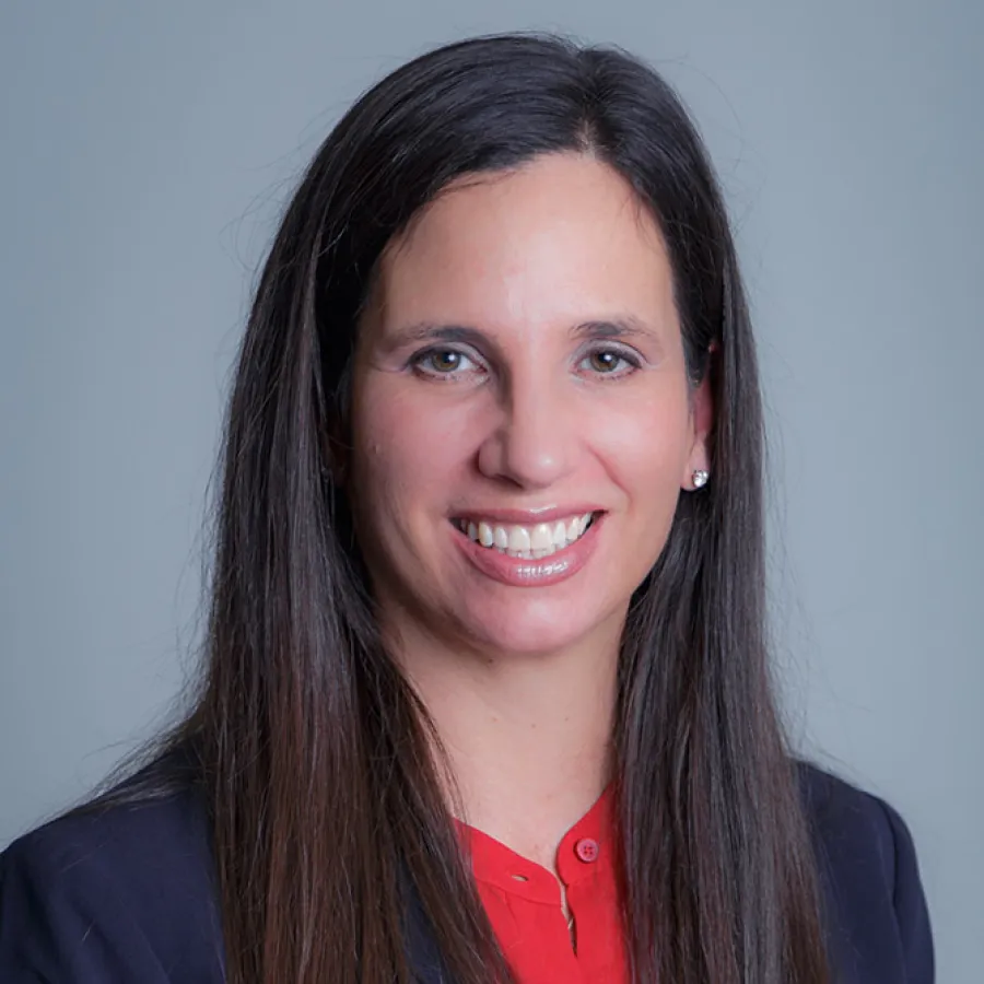Professional headshot of a smiling woman with long dark hair wearing a red blouse and dark blazer against gray background