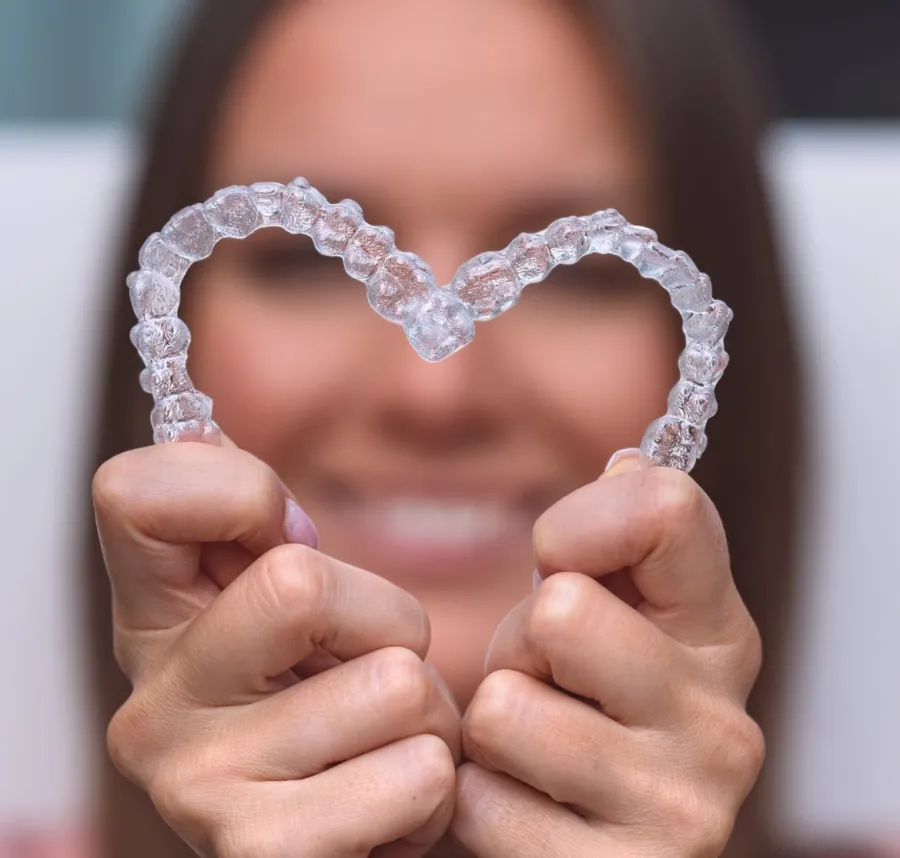 Close-up of hands holding two clear dental aligners shaped into a heart with a smiling woman blurred in the background
