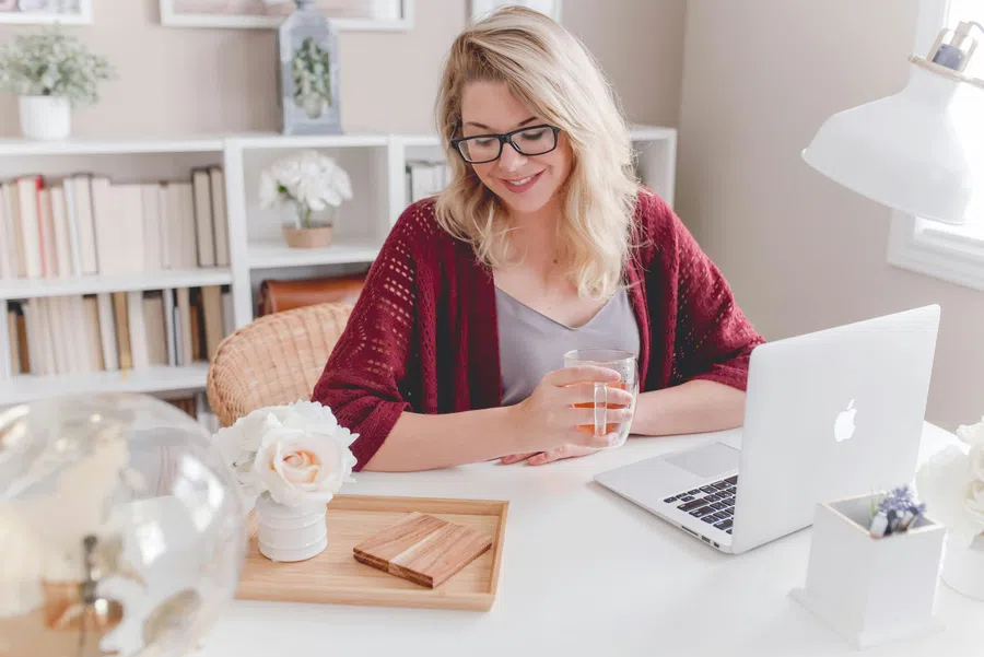 Smiling woman with glasses drinking water while sitting at desk with laptop in cozy home office setting