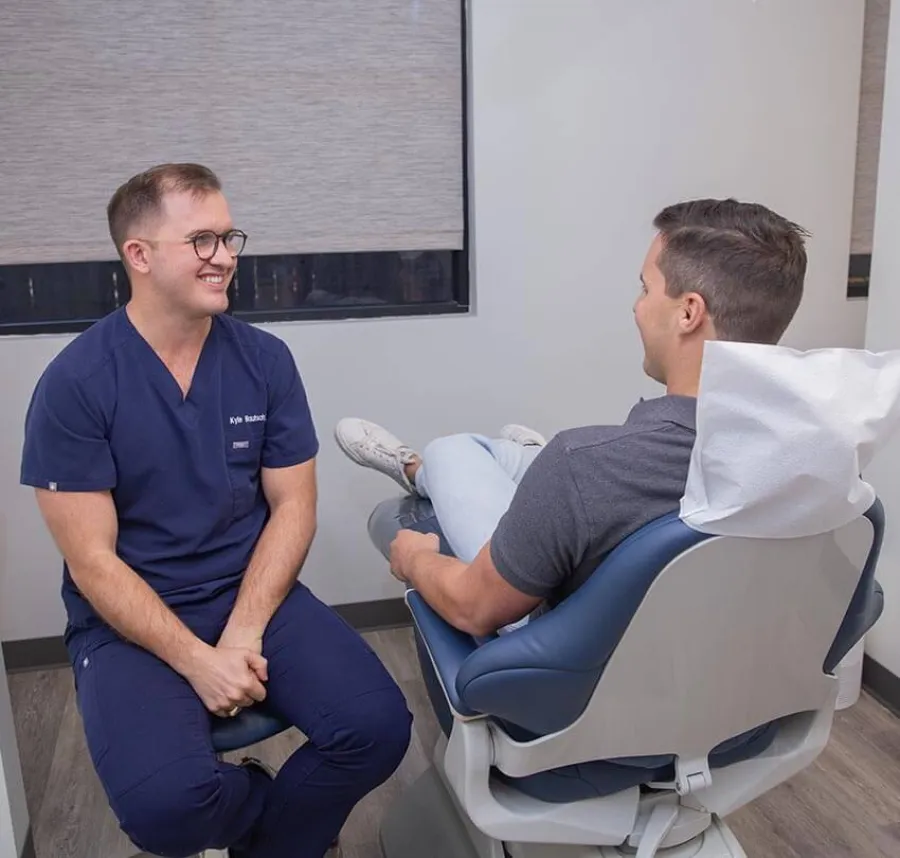 Dentist in navy scrubs smiling and talking to a patient sitting in a dental chair in a clinic room.