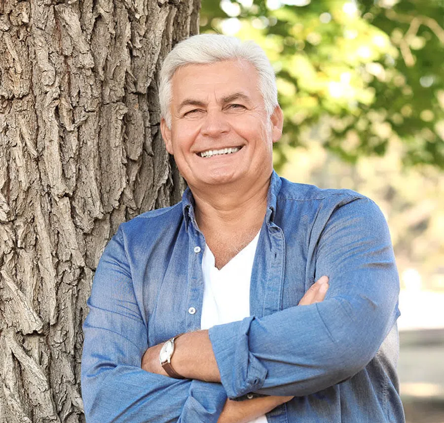 Smiling older man in blue shirt leaning against a tree outdoors with crossed arms and green foliage background