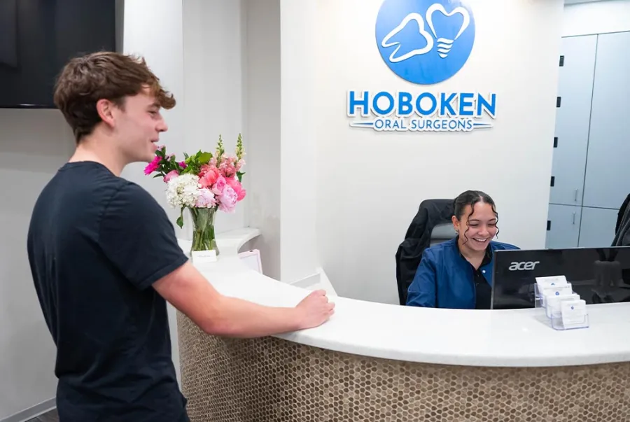 Young male patient speaking with a smiling receptionist at Hoboken Oral Surgeons front desk with flowers in background