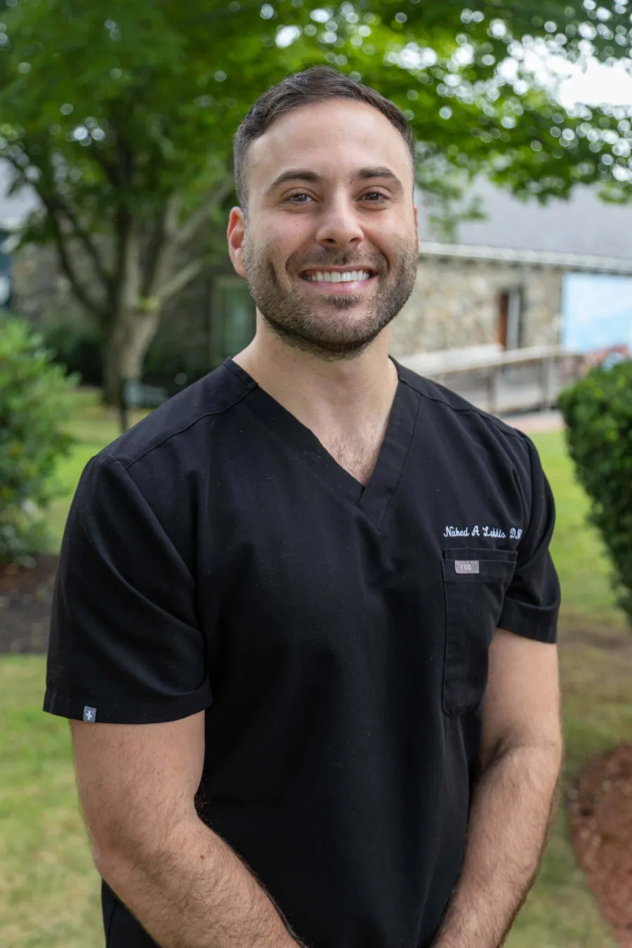 Smiling man in black medical scrubs standing outdoors with trees and stone building in the background.