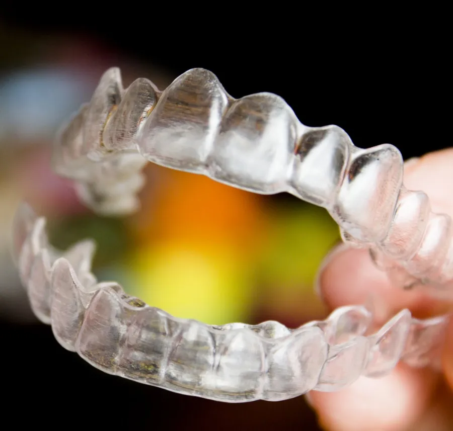 Close-up of a clear dental aligner held between fingers with a blurred colorful background.