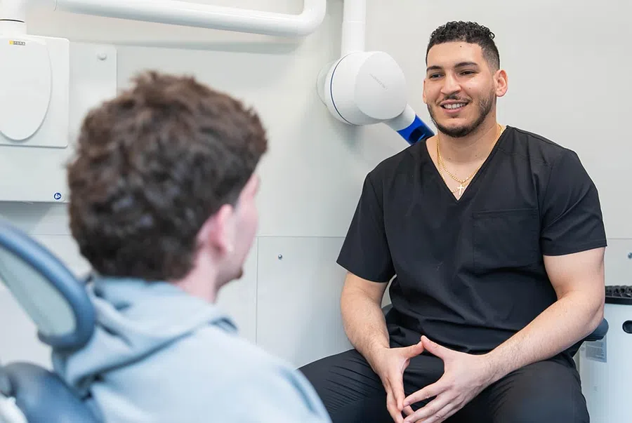 Dentist in black scrubs talking to a patient seated in a dental chair in a modern clinic.