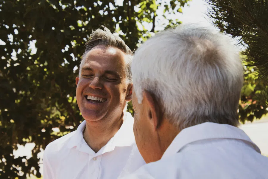 Two men outdoors in white shirts laughing and enjoying a sunny day with leafy trees in the background