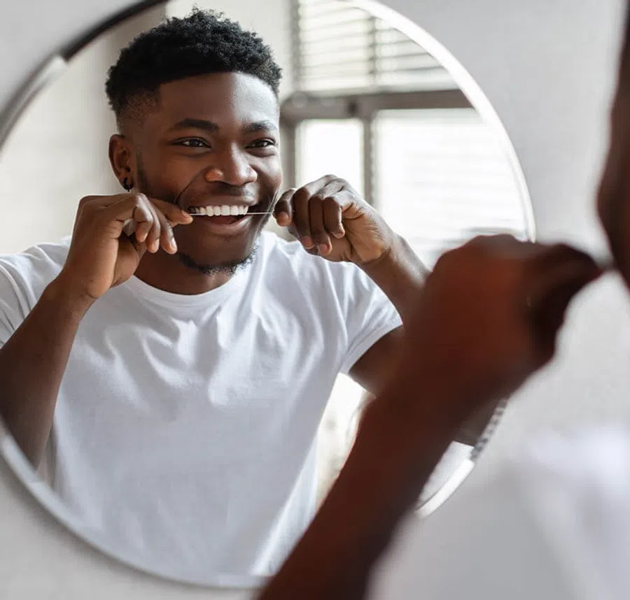 Smiling young man flossing teeth in front of a round bathroom mirror wearing a white t-shirt.