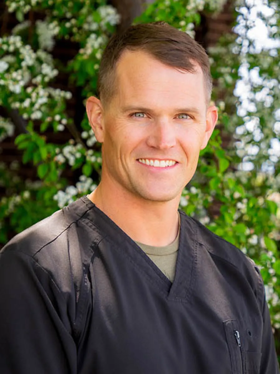 Smiling man in black medical scrubs standing outdoors with green foliage and white flowers in background