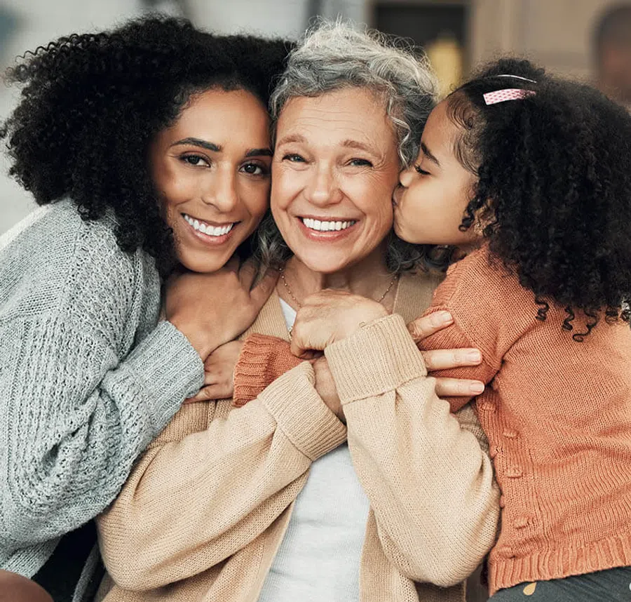 Three generations of women smiling and embracing, showing love and family bond indoors.