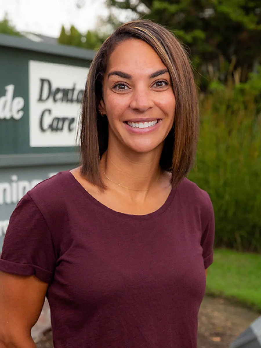 Smiling woman with shoulder-length hair wearing a maroon shirt standing outdoors near a dental care sign.