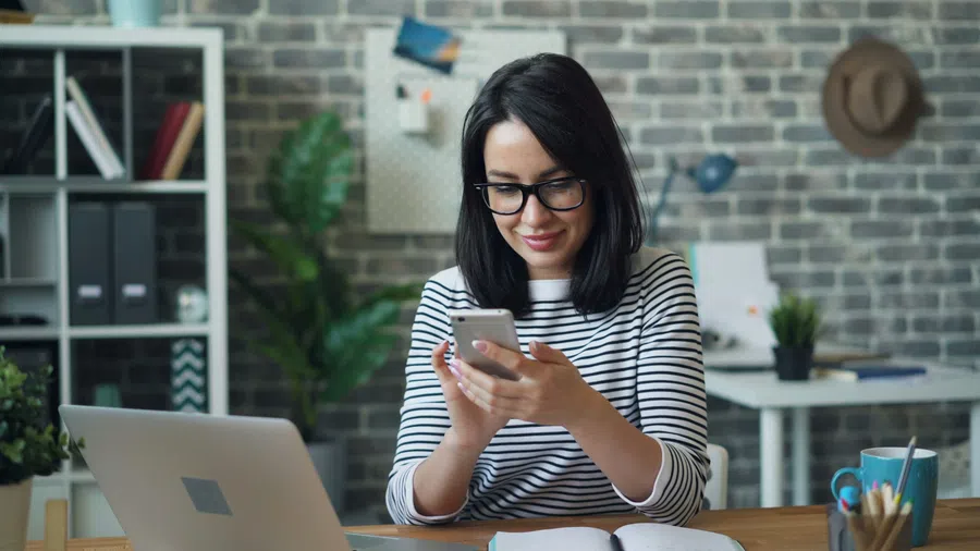 Woman in orange shirt smiling and talking while working on laptop during a meeting in bright office.