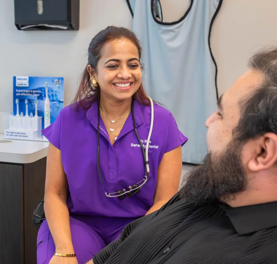 Smiling female dentist in purple scrubs talking with male patient in dental office setting.