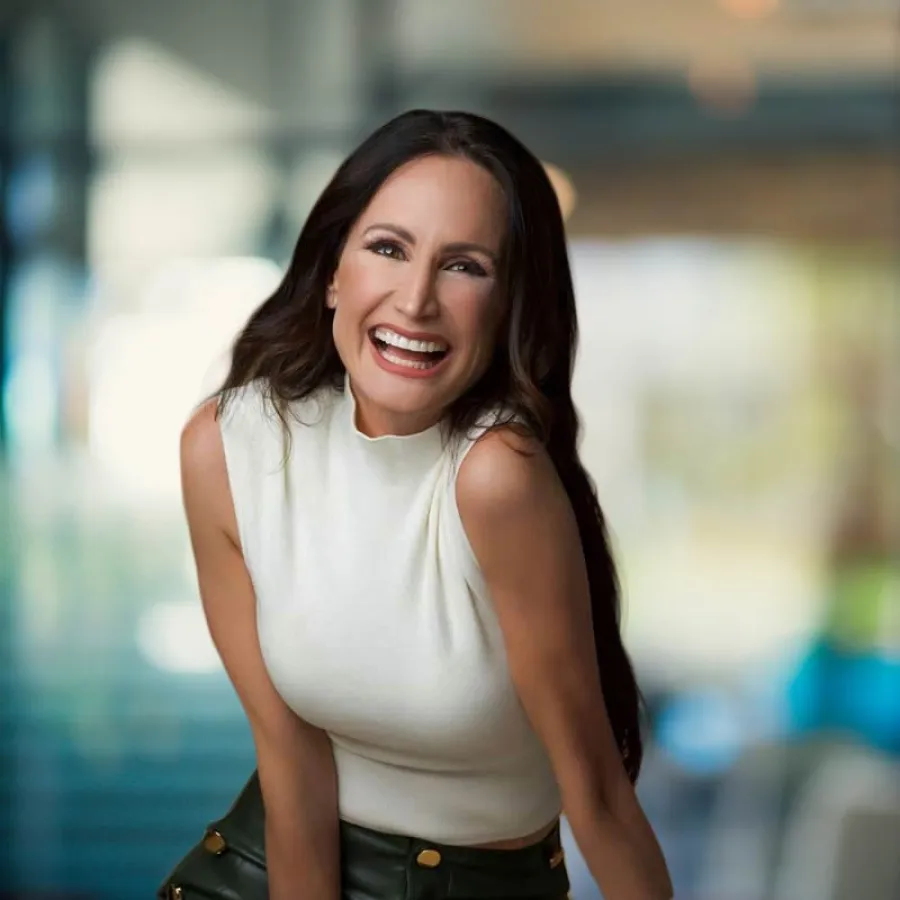 Smiling woman with long dark hair wearing a white sleeveless top and green pants against a blurred background