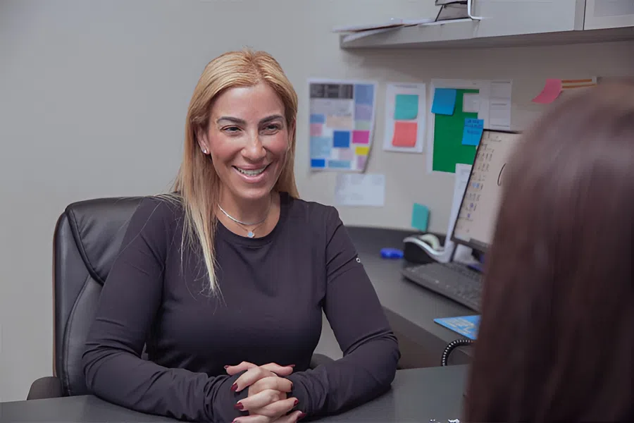Smiling woman in black shirt sitting at desk facing another person in office with computer and notes.