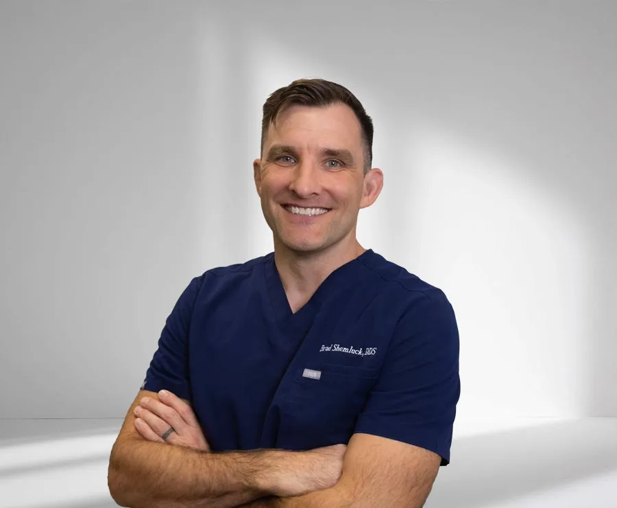 Smiling male dentist in navy blue scrubs with arms crossed against a plain light background.