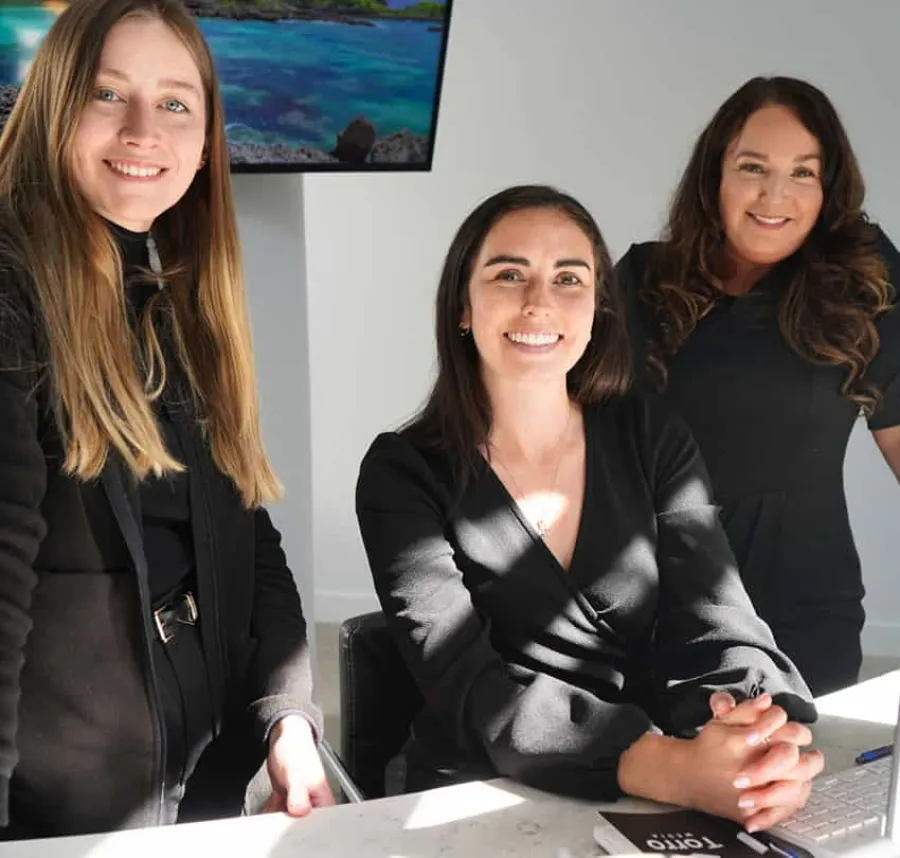 Three professional women in black attire smiling in a bright office environment with a computer and notebook.