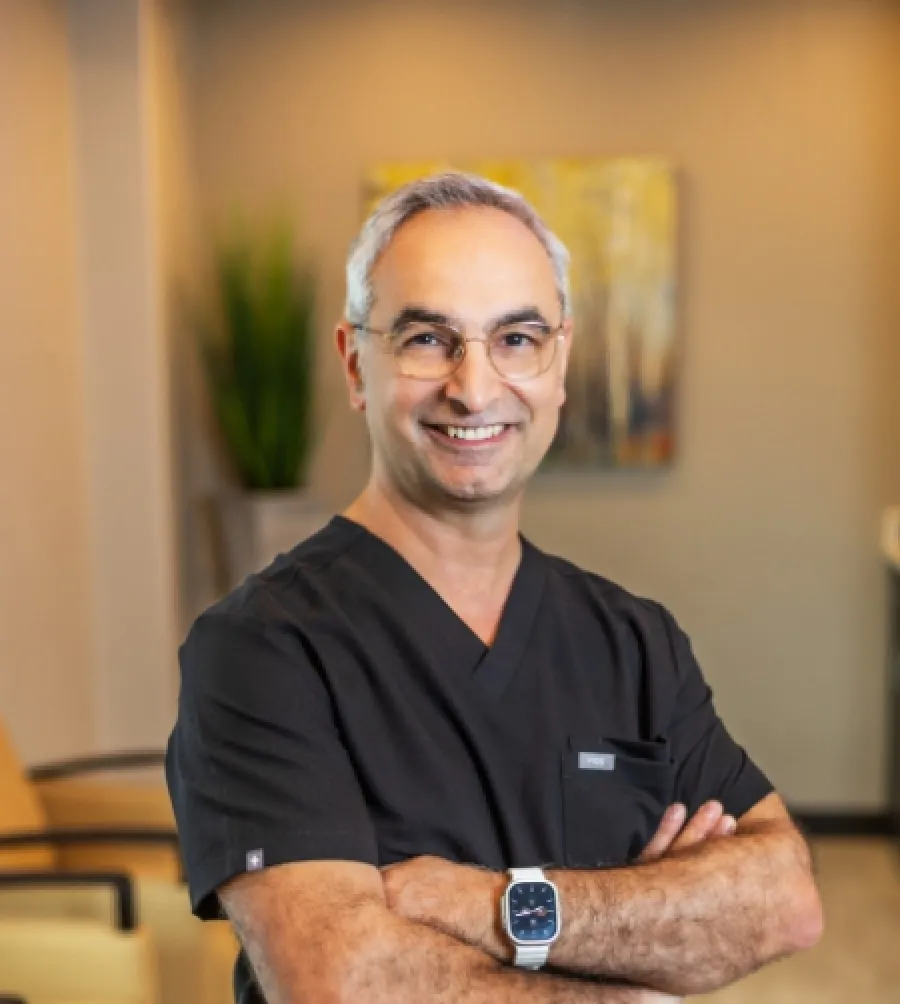 Smiling middle-aged male doctor with glasses and smartwatch wearing black scrubs, standing with arms crossed indoors.