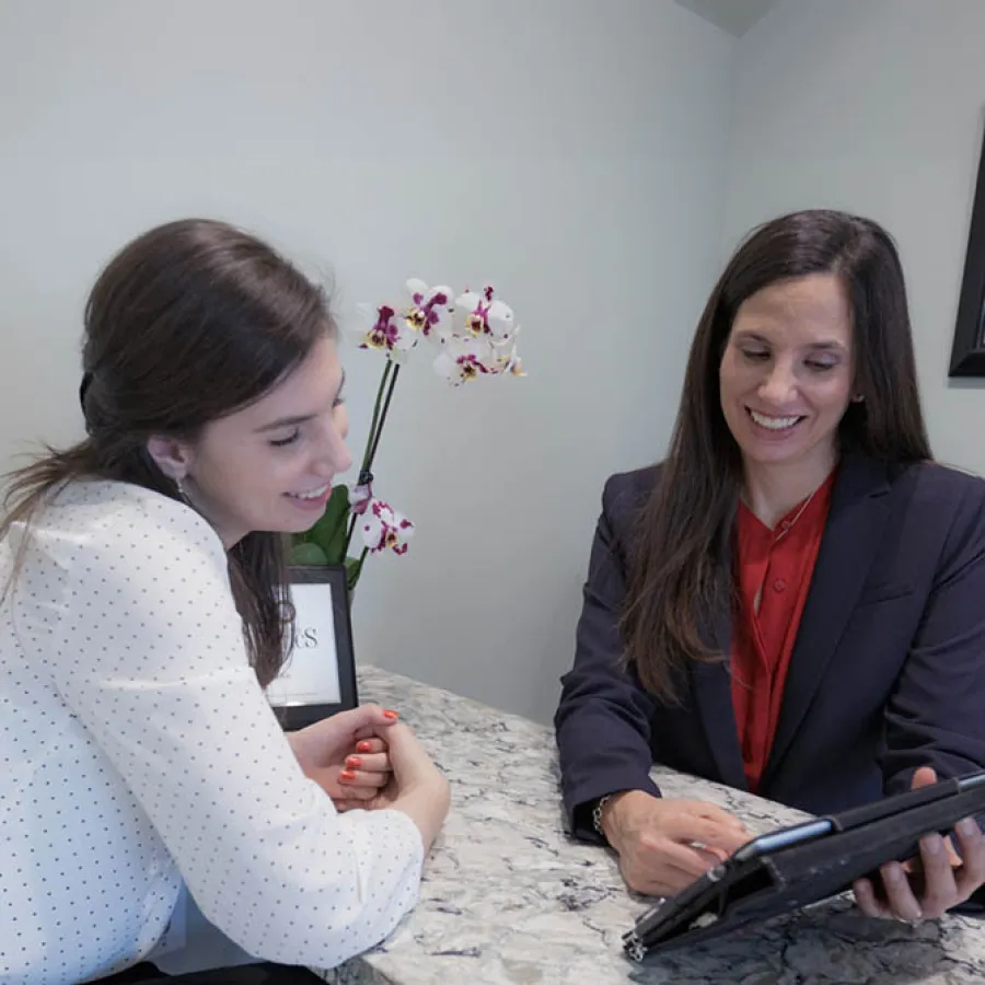 Two women smiling and discussing while viewing a tablet at a marble table with orchid flowers in the background