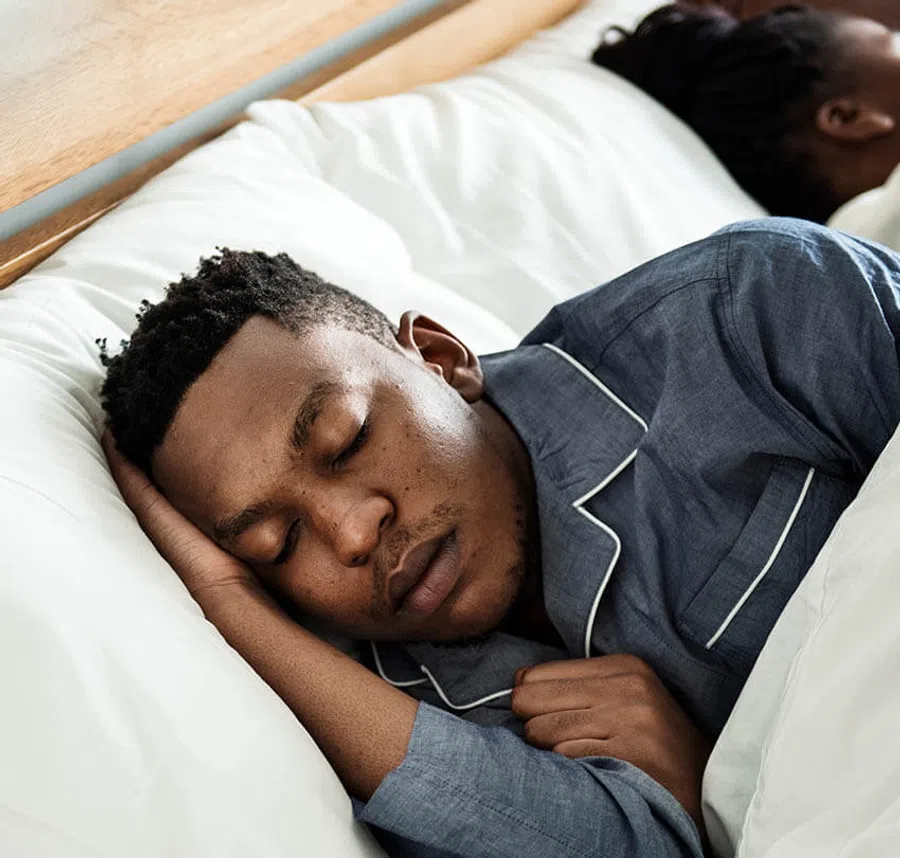 Man peacefully sleeping in blue pajamas on a white bed with wooden headboard, resting head on hand.