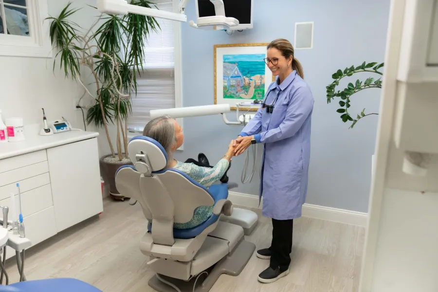 Dentist in a blue coat shaking hands with a patient seated in a dental chair in a modern clinic.