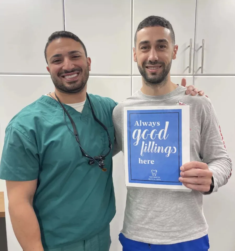 Smiling dentist in scrubs with patient holding a sign reading Always good fillings here.