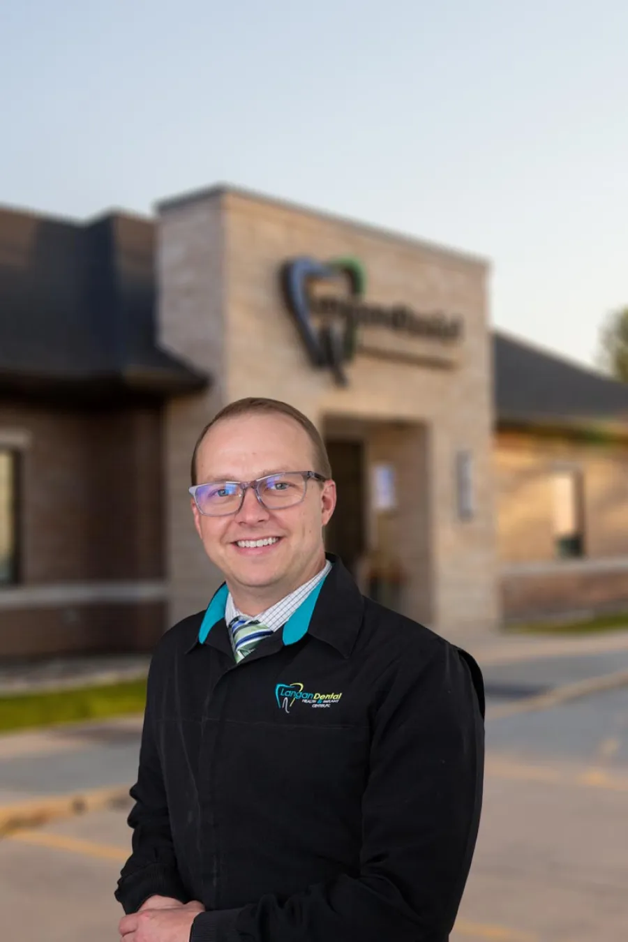 Smiling man in glasses wearing a Langan Dental jacket stands outside the dental office building.