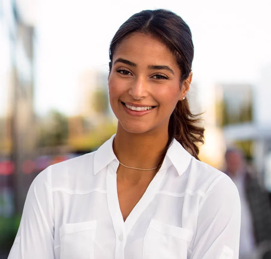 Smiling young woman with dark hair in a white shirt standing outdoors in soft focus background.