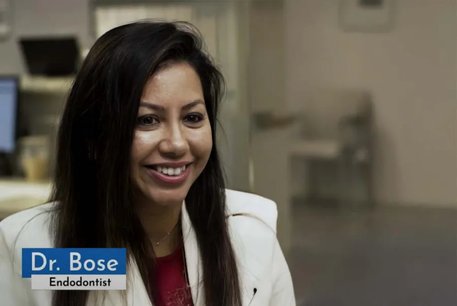 Smiling female endodontist Dr. Bose in a white coat at a dental office with blurred background.