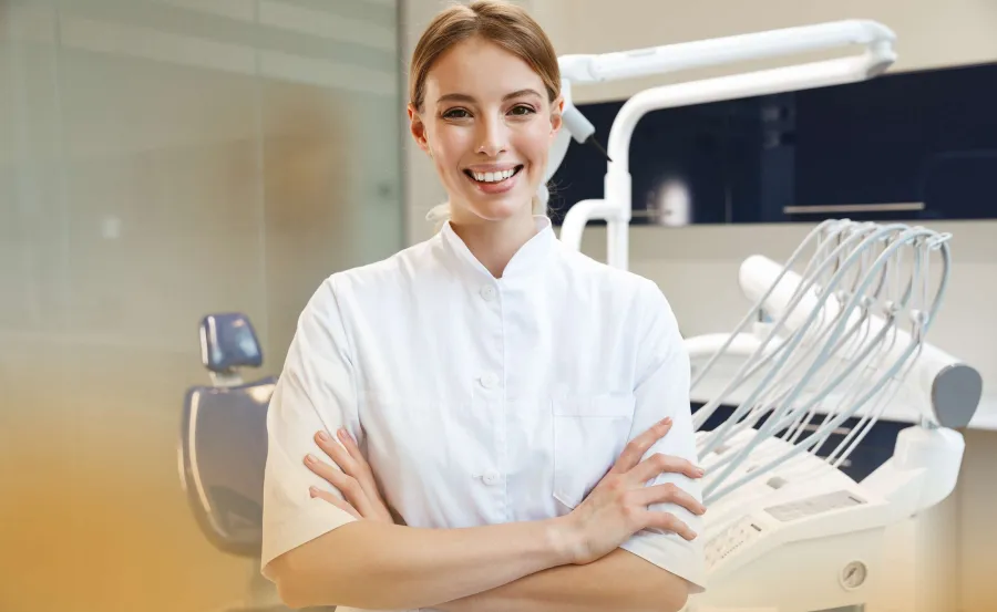 Smiling female dentist in white coat stands confidently in modern dental clinic with chair and equipment.