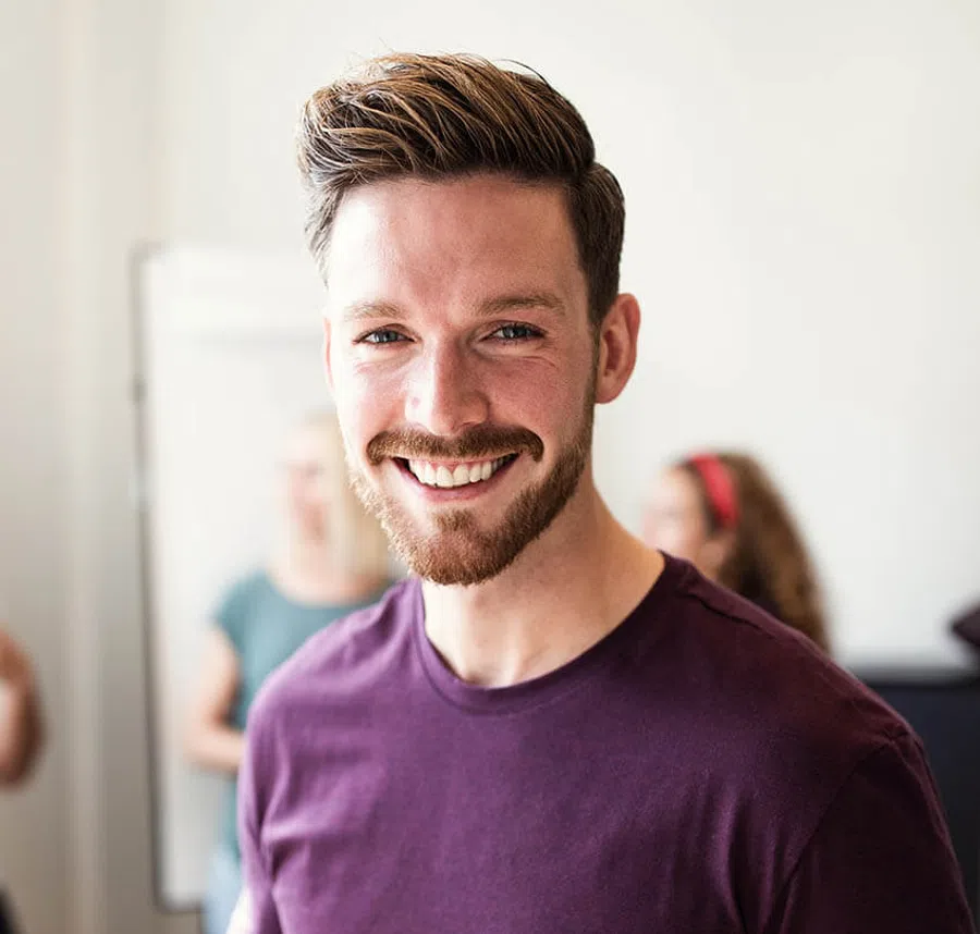 Smiling young man with beard and purple shirt in a bright office setting with blurred people in background