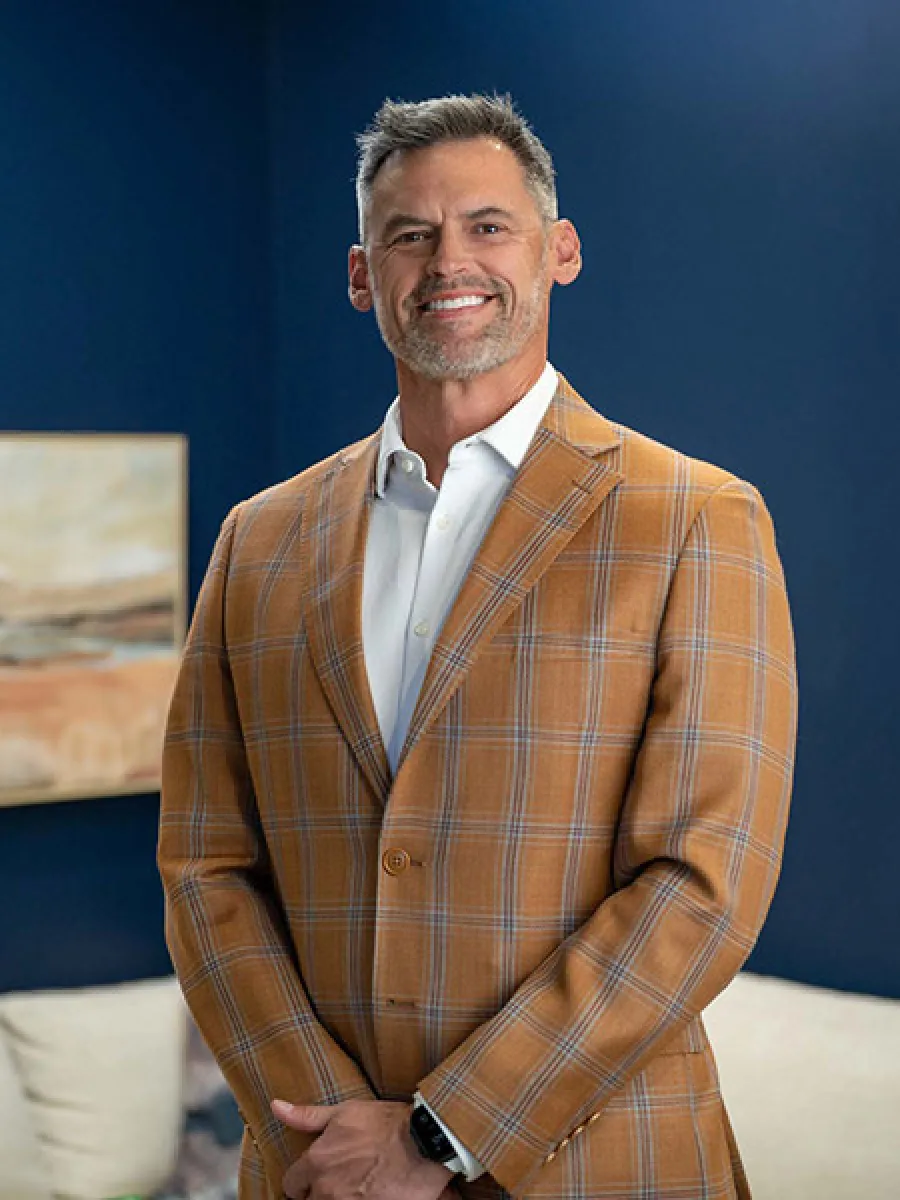 Smiling man in brown plaid blazer and white shirt stands against a blue wall with abstract art and cushions nearby.