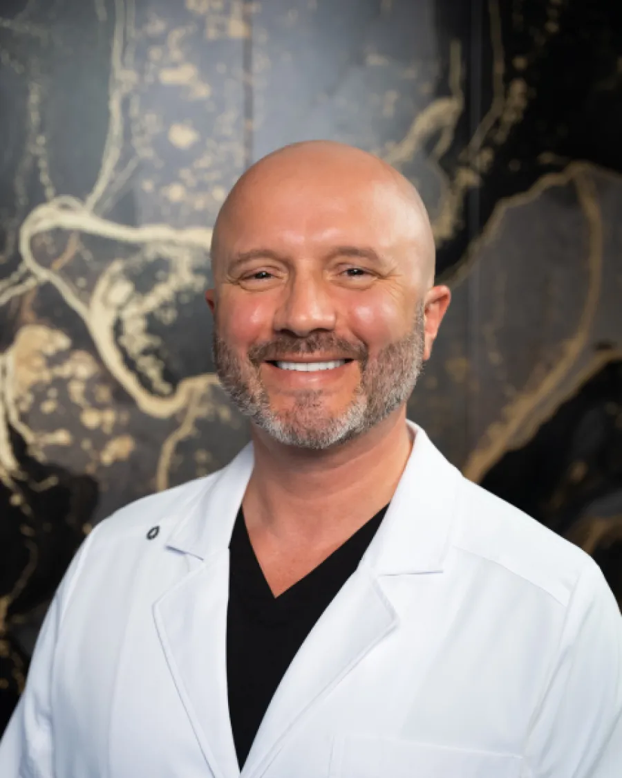 Smiling bald man with beard wearing white lab coat and black shirt against artistic dark background