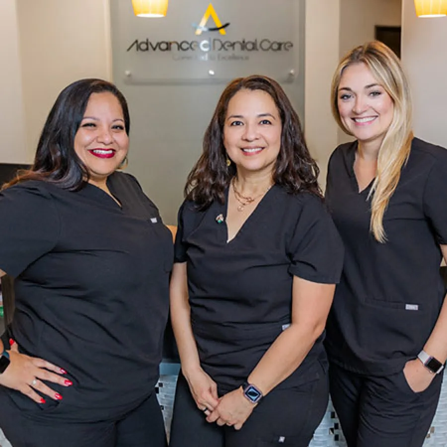 Three female dental professionals in black scrubs smiling at Advance Dental Care clinic reception area.