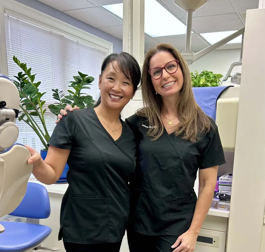 Two female dental professionals smiling in a bright dental clinic wearing black scrubs with plants in the background