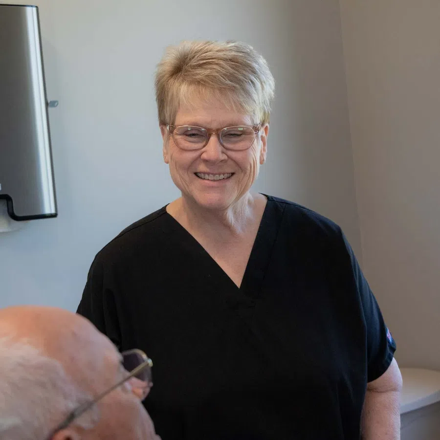 Smiling healthcare professional in black scrubs standing in a medical office with elderly patient sitting nearby.