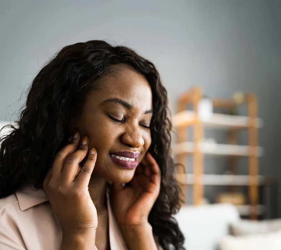 Woman with closed eyes touching her jaw in pain, likely experiencing toothache or jaw discomfort indoors.