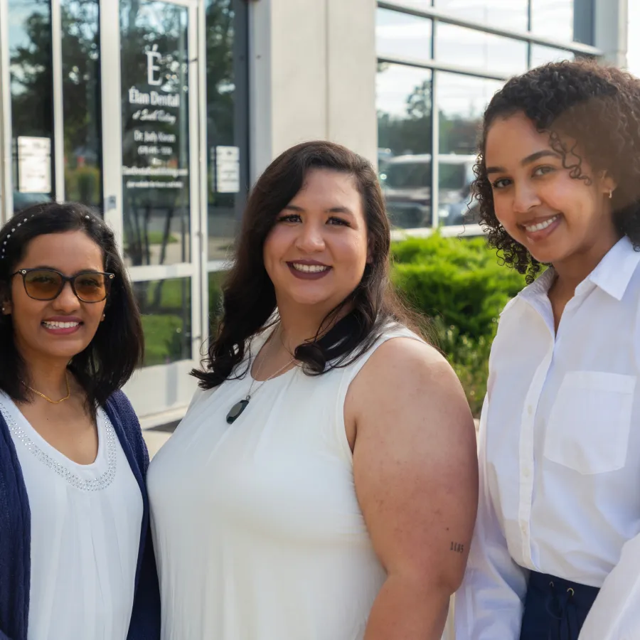 Three diverse women smiling outside a dental office on a sunny day with greenery and glass windows.
